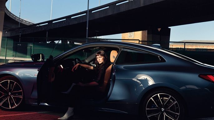 Imagine cu partea laterală a BMW Seria 4 Coupé, G22, 2020, cu jantele din aliaj uşor Imagine cu partea laterală a BMW Seria 4 Coupé, G22, 2020, în Albastru Arctic Race metalizat, cu jantele din aliaj uşor şi un model stând în automobil, cu portiera deschisă