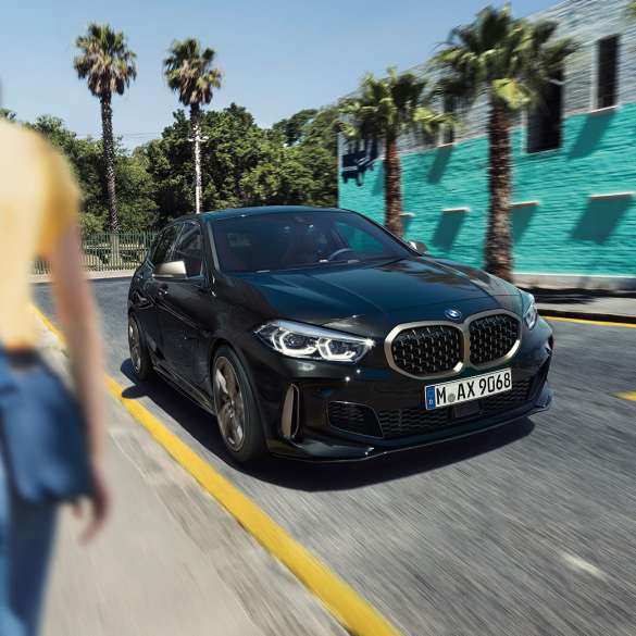 Imagine cu trei sferturi din partea frontală a BMW M135i xDrive. Imagine cu trei sferturi din partea frontală a BMW M135i xDrive în Negru Sapphire metalizat, rulând, cu o femeie în prim-plan.