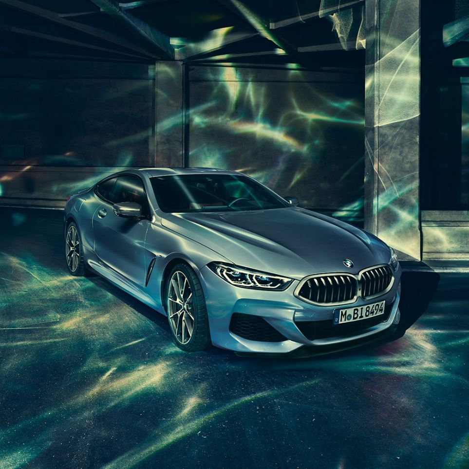Imagine cu trei sferturi din partea frontală stânga a BMW M850i xDrive Coupé. Imagine cu trei sferturi din partea frontală stânga a BMW M850i xDrive Coupé în Albastru Barcelona metalizat, staţionând în faţa unei clădiri.
