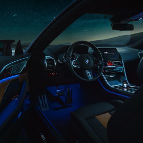 Interiorul BMW M8 Competition Coupé. Imagine cu interiorul BMW M8 Competition Coupé, cu volan M îmbrăcat în piele şi lumină ambientală albastră, pe timp de noapte.