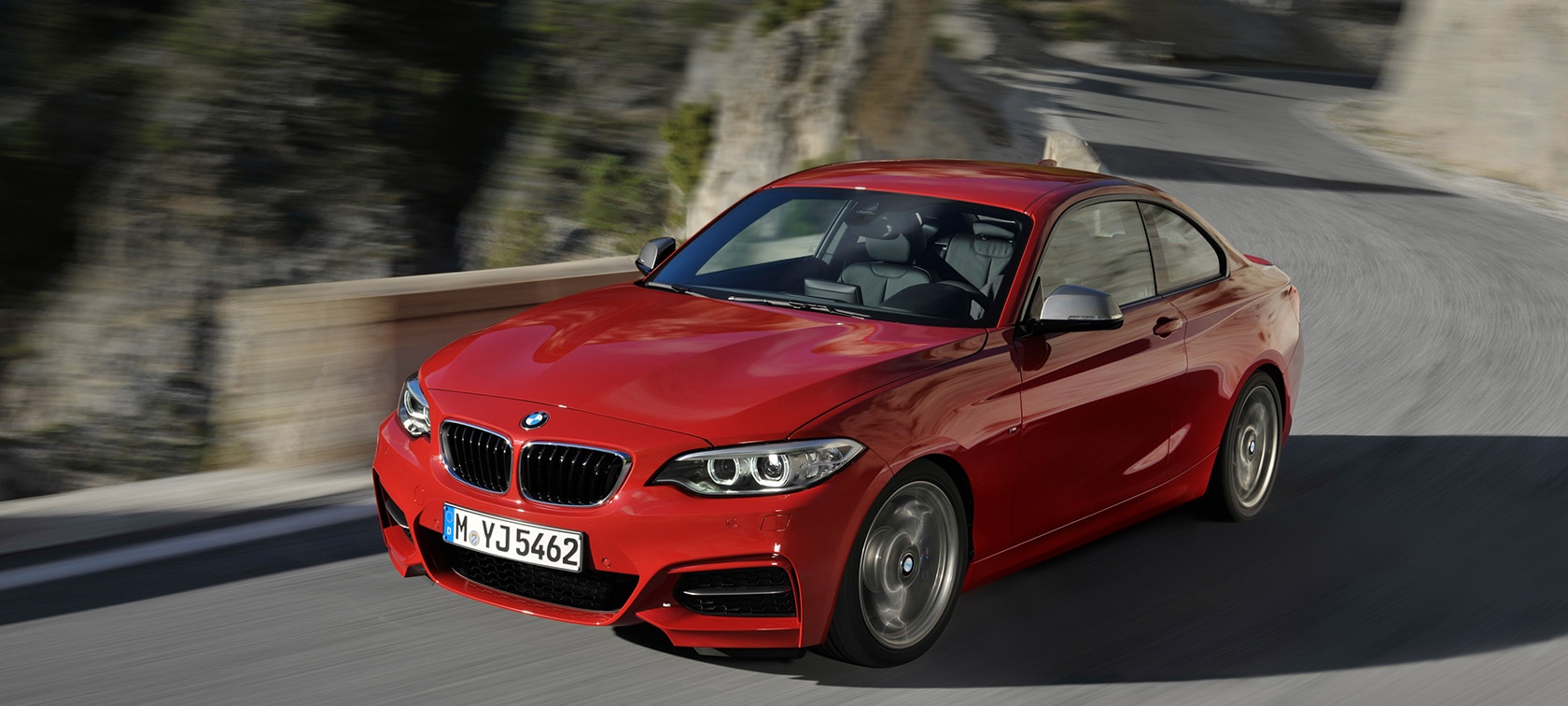 BMW Seria 2, imagine cu trei sferturi din partea frontală, rulând pe drum în faţa unui peisaj BMW Seria 2, imagine cu trei sferturi din partea frontală a BMW, rulând pe drum în faţa unui peisaj