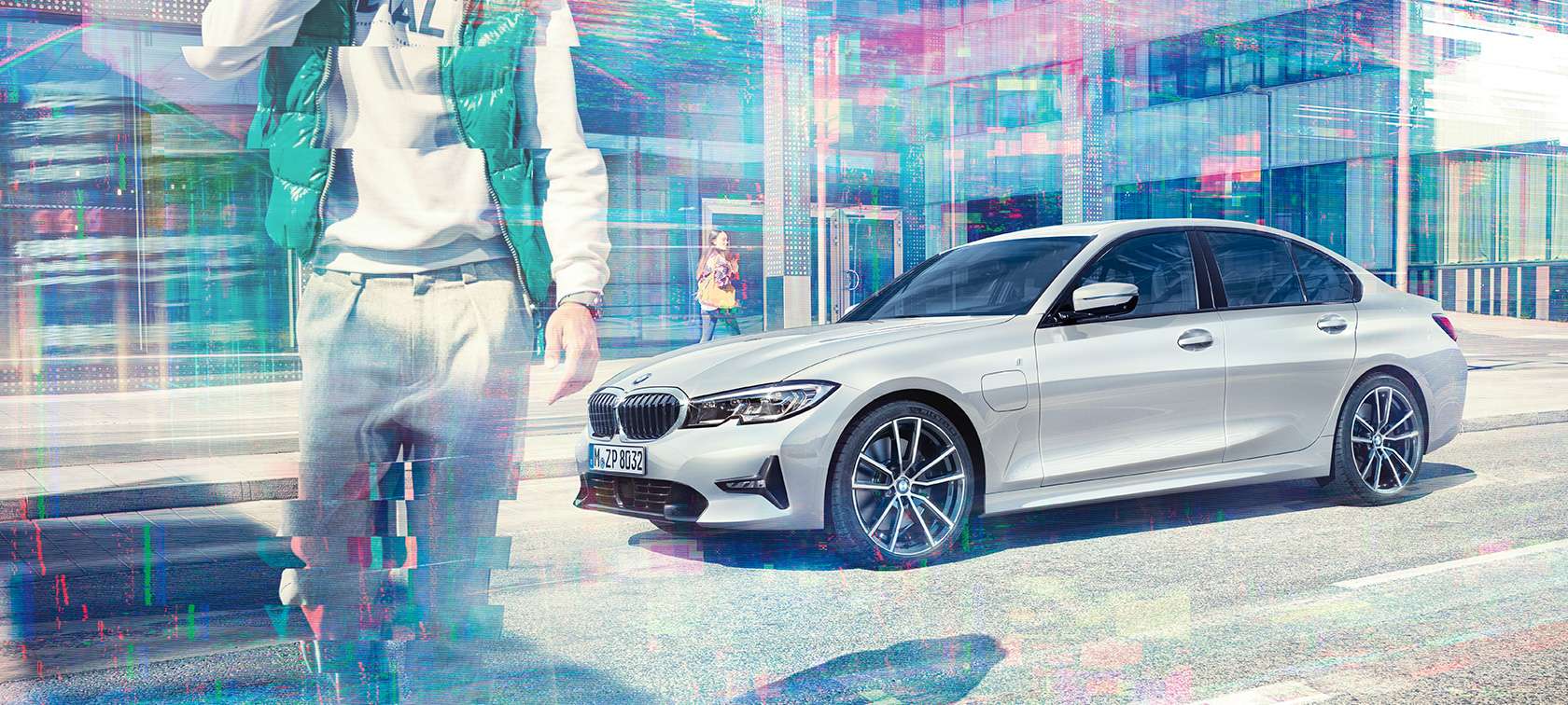 Imagine din lateral cu BMW 330e al BMW Seria 3 Sedan. Imagine cu trei sferturi din partea laterală a BMW 330e Sedan cu pasageri în mers, în oraş, aflat în faţa unei clădiri din sticlă.