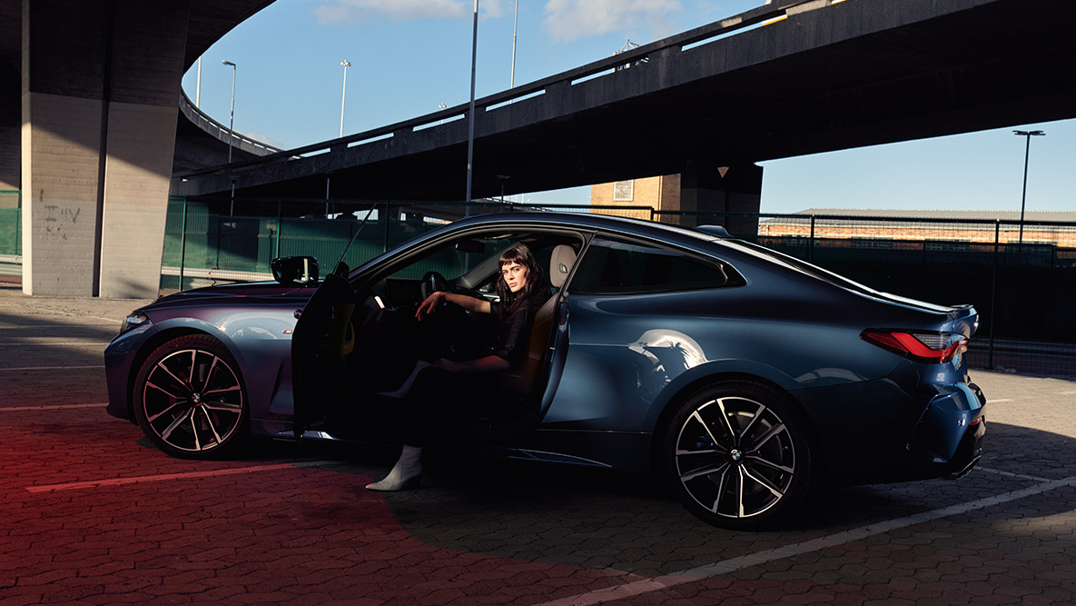 Imagine cu partea laterală a BMW Seria 4 Coupé, G22, 2020, cu jantele din aliaj uşor Imagine cu partea laterală a BMW Seria 4 Coupé, G22, 2020, în Albastru Arctic Race metalizat, cu jantele din aliaj uşor şi un model stând în automobil, cu portiera deschisă