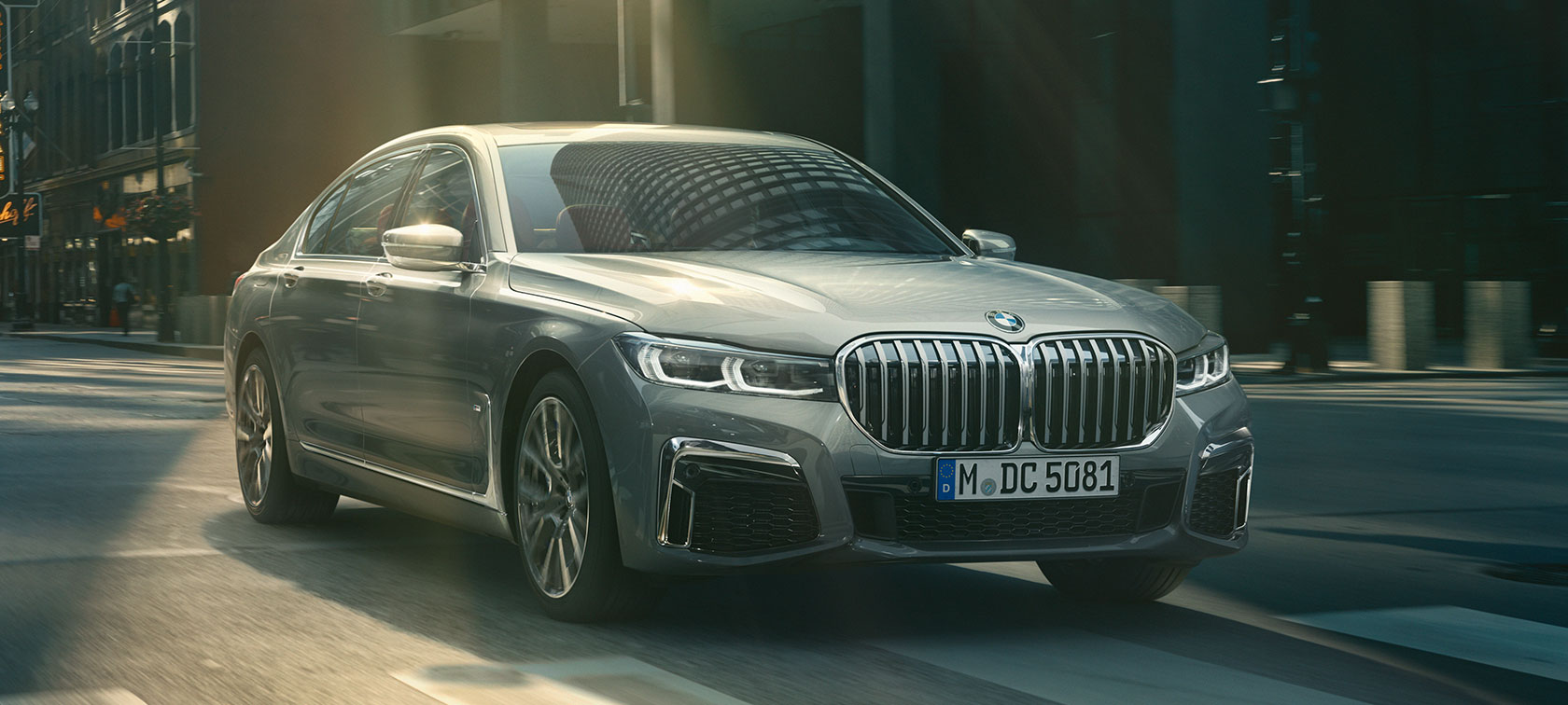 Imagine cu trei sferturi din partea frontală a unui BMW Seria 7 Sedan Imagine cu trei sferturi din partea frontală a unui BMW Seria 7 Sedan gri rulând într-un peisaj urban