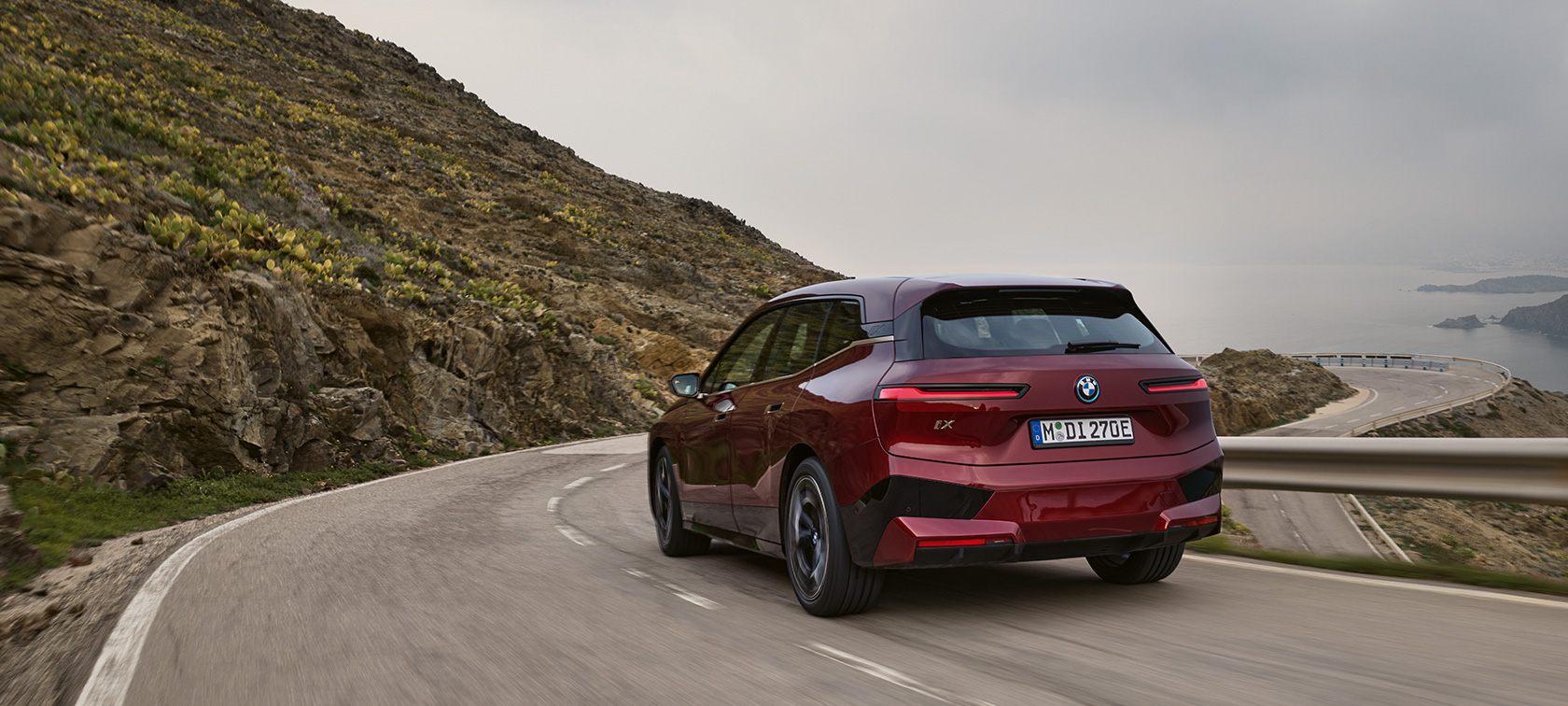 BMW iX i20 2021 SUV electric BMW iX xDrive50 în timp ce se deplasează pe o şosea de coastă BMW iX i20 2021 SUV electric BMW iX xDrive50 Roşu Aventurin Imagine din partea din spate în timp ce se deplasează pe o şosea de coastă