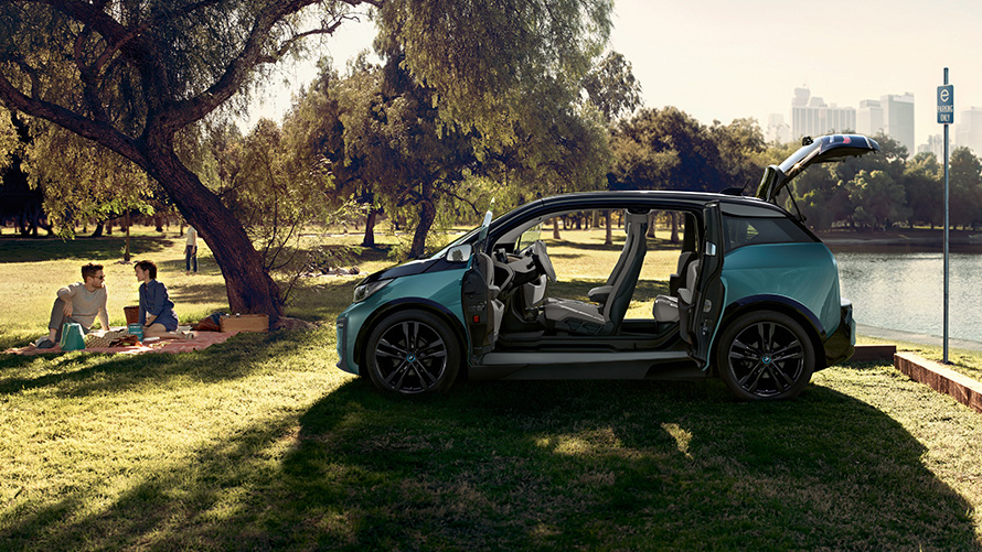 Imagine cu BMW i3, 2018, în parc Imagine cu partea laterală a BMW i3, I01, 2018, în Bej Jucaro cu detalii în Gri Frozen metalizat, cu portierele deschise într-un parc
