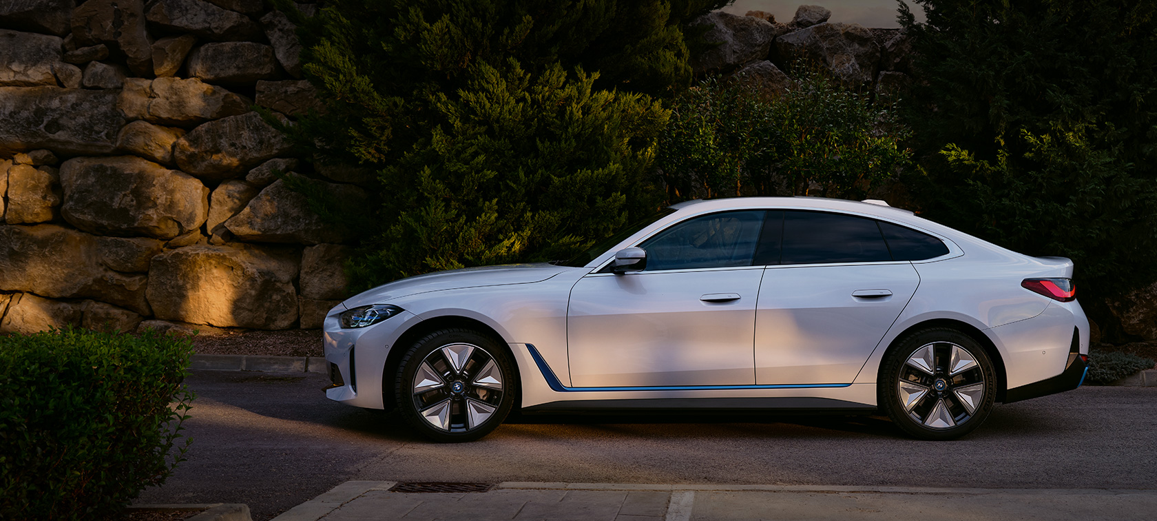 Imagine cu trei sferturi din partea frontală a unui BMW i4 eDrive40 G26 2021 Alb Mineral staţionând în faţa unui lac cu un deal în fundal