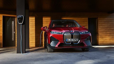 Imagine din partea frontală a unui BMW iX staţionând lângă o staţie de încărcare Imagine din partea frontală a unui BMW Seria i BMW iX staţionând lângă o staţie de încărcare