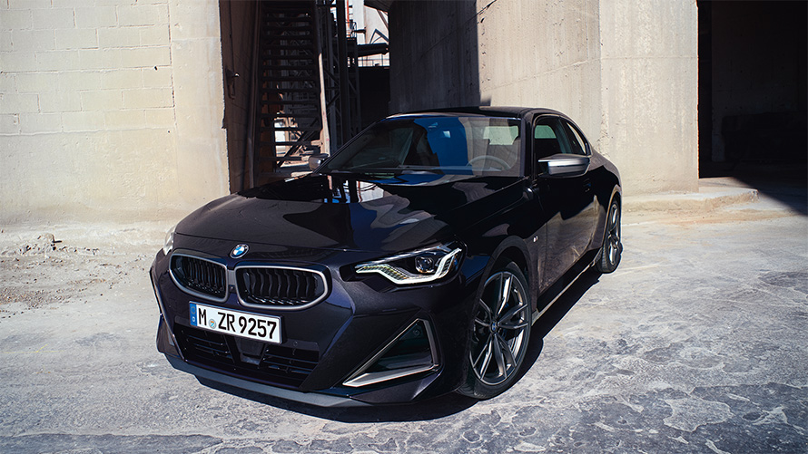 Imagine cu trei sferturi din partea frontală a BMW M240i xDrive Coupé G42 2021, staţionând Imagine cu trei sferturi din partea frontală a BMW M240i xDrive Coupé G42 2021 în Negru Sapphire, staţionând în faţa unui depozit