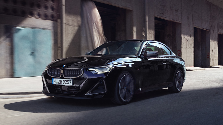 Imagine cu trei sferturi din partea laterală a BMW M240i xDrive Coupé G42 2021, rulând Imagine cu trei sferturi din partea laterală a BMW M240i xDrive Coupé G42 2021 în Negru Sapphire, rulând în faţa unor depozite