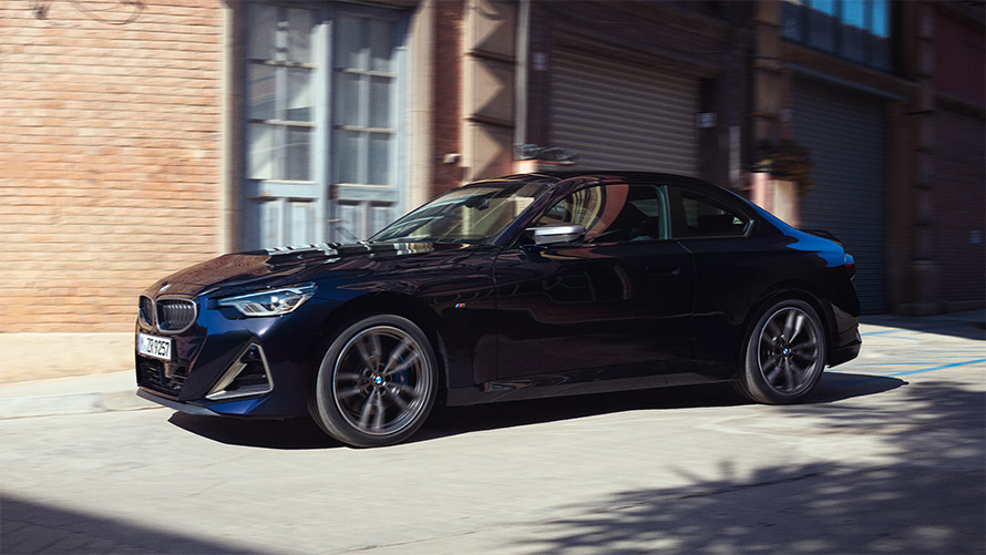Imagine din partea laterală cu BMW M240i xDrive Coupé G42 2021, staţionând Imagine din partea laterală BMW M240i xDrive Coupé G42 2021 în Negru Sapphire, staţionând în faţa unui depozit