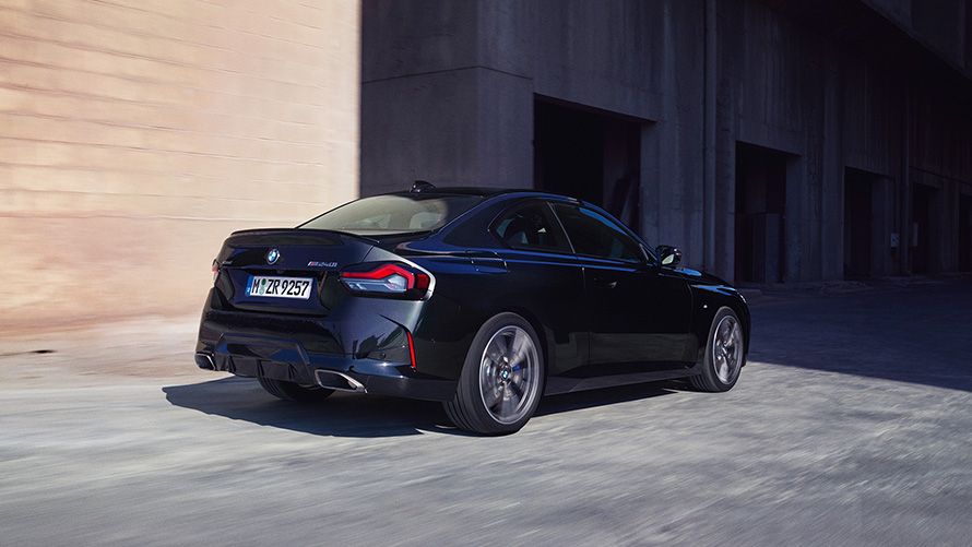 Imagine cu trei sferturi din partea din spate a BMW M240i xDrive Coupé G42 2021, staţionând Imagine cu trei sferturi din partea din spate cu BMW M240i xDrive Coupé G42 2021, în Negru Sapphire, staţionând în faţa unui depozit