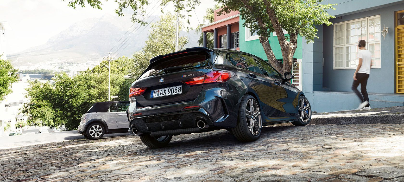 Imagine cu trei sferturi din partea spate a BMW M135i xDrive. Imagine cu trei sferturi din partea spate a BMW M135i xDrive în Negru Sapphire metalizat, parcând, în faţa unei clădiri rezidenţiale.