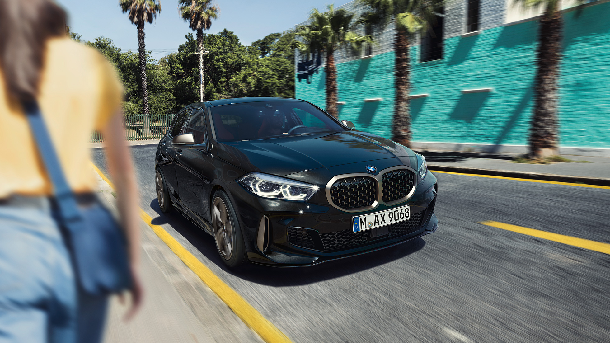 Imagine cu trei sferturi din partea frontală a BMW M135i xDrive. Imagine cu trei sferturi din partea frontală a BMW M135i xDrive în Negru Sapphire metalizat, rulând, cu o femeie în prim-plan.