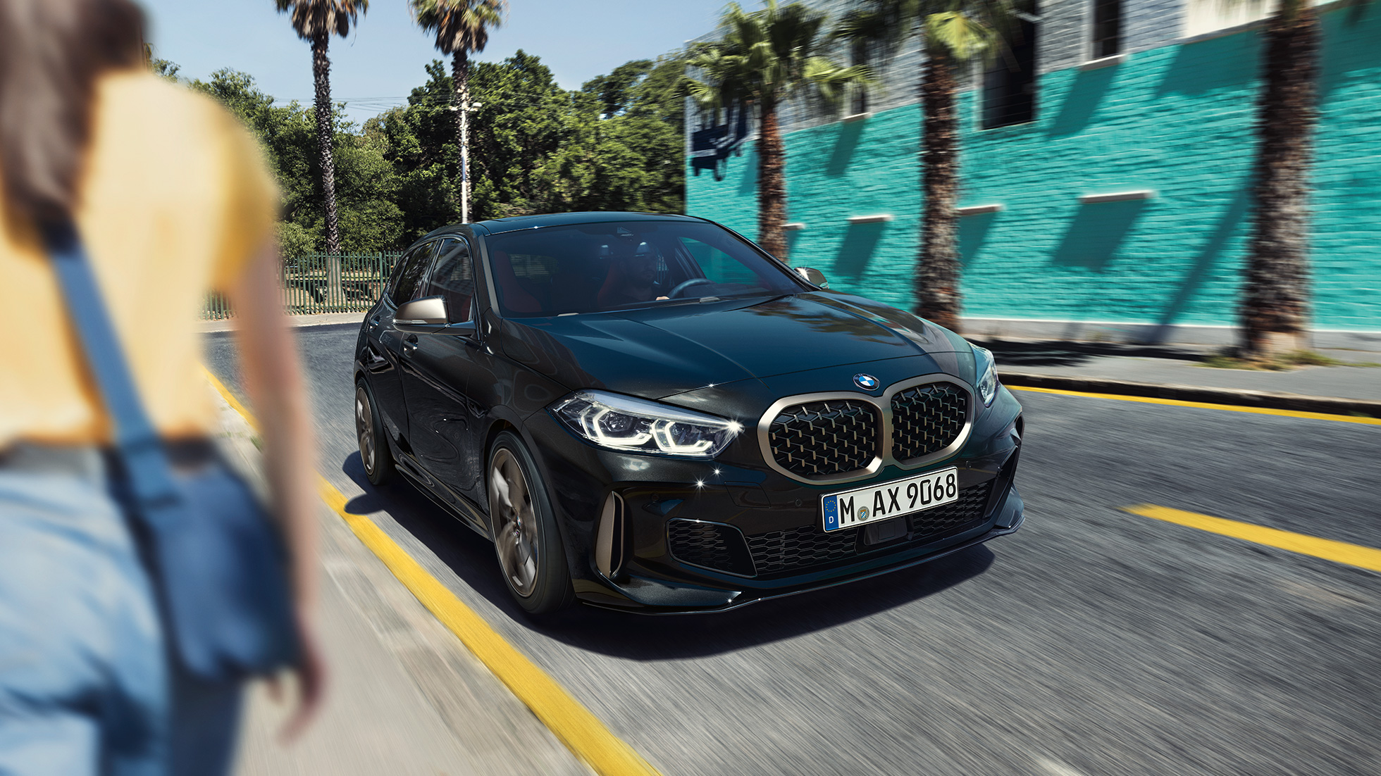 Imagine cu trei sferturi din partea frontală a BMW M135i xDrive. Imagine cu trei sferturi din partea frontală a BMW M135i xDrive în Negru Sapphire metalizat, rulând, cu o femeie în prim-plan.