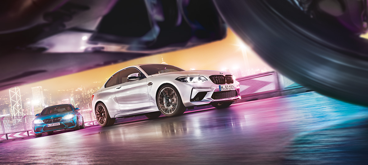 BMW M2 Competition pe asfalt reflectorizant Două automobile BMW M2 Competition pe un circuit de curse în faţa unui peisaj urban nocturn, vedere din stradă.