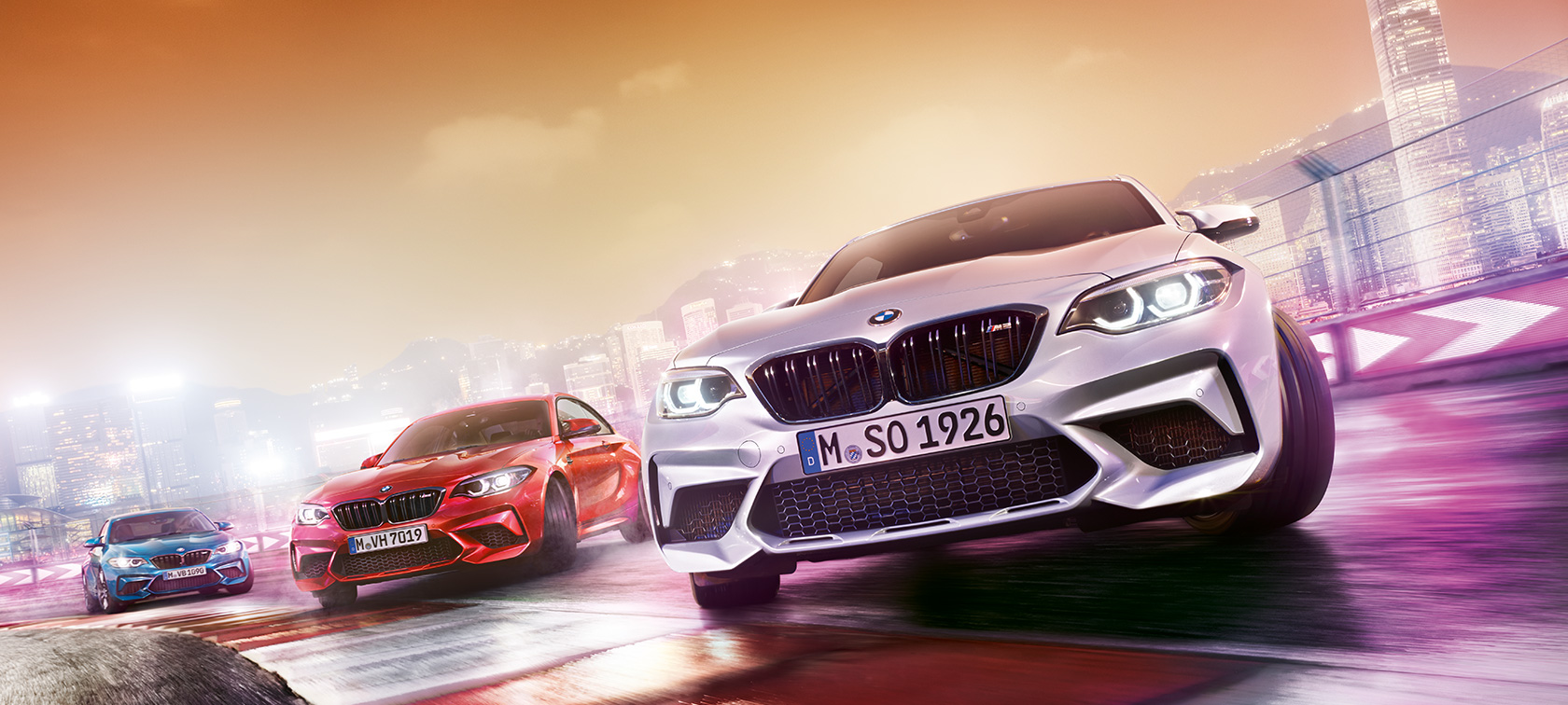 BMW M2 Competition în acţiune Trei automobile BMW M2 Competition fac drifting pe un circuit de curse în faţa unui peisaj urban nocturn.