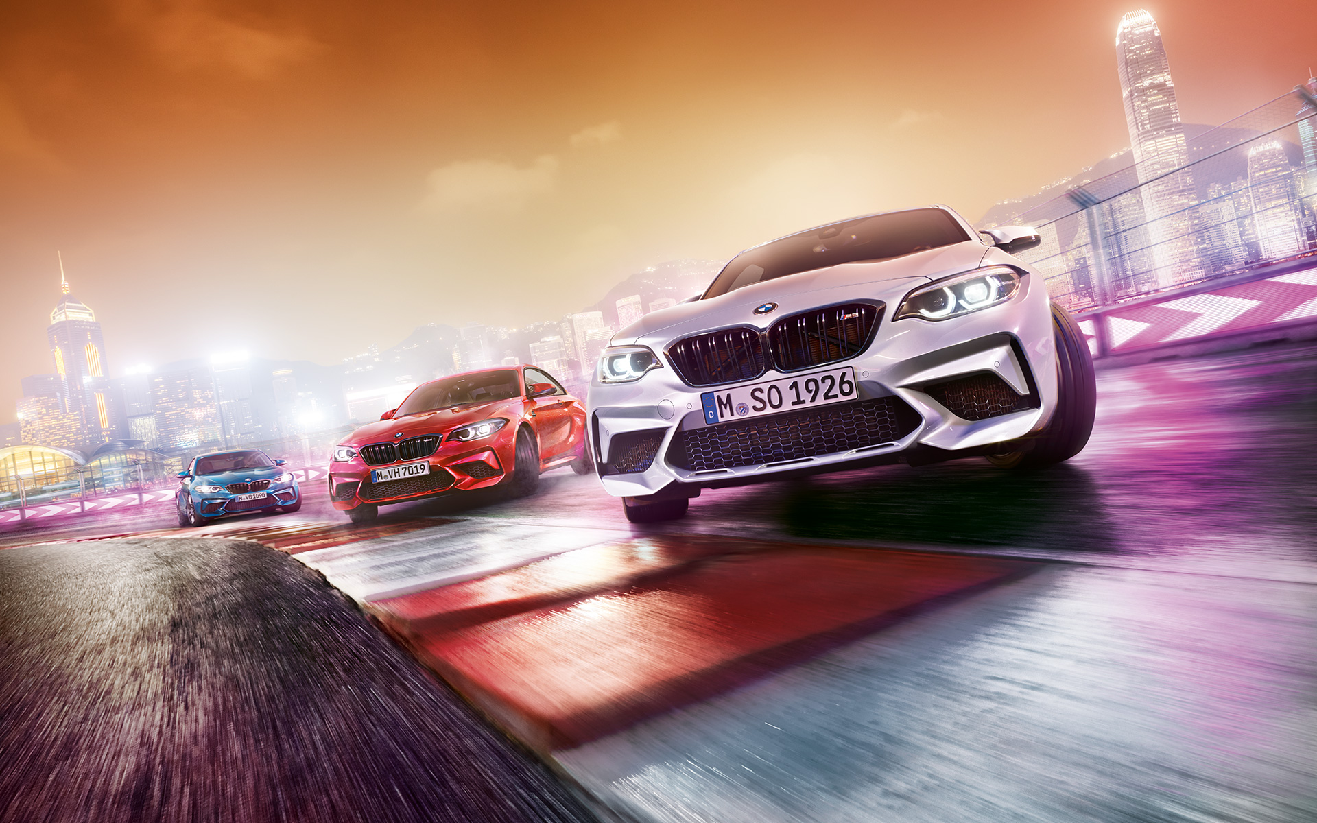 BMW M2 Competition în acţiune Trei automobile BMW M2 Competition fac drifting pe un circuit de curse în faţa unui peisaj urban nocturn.