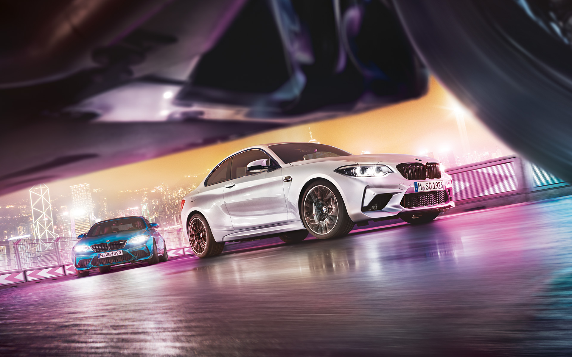 BMW M2 Competition pe asfalt reflectorizant Două automobile BMW M2 Competition pe un circuit de curse în faţa unui peisaj urban nocturn, vedere din stradă.