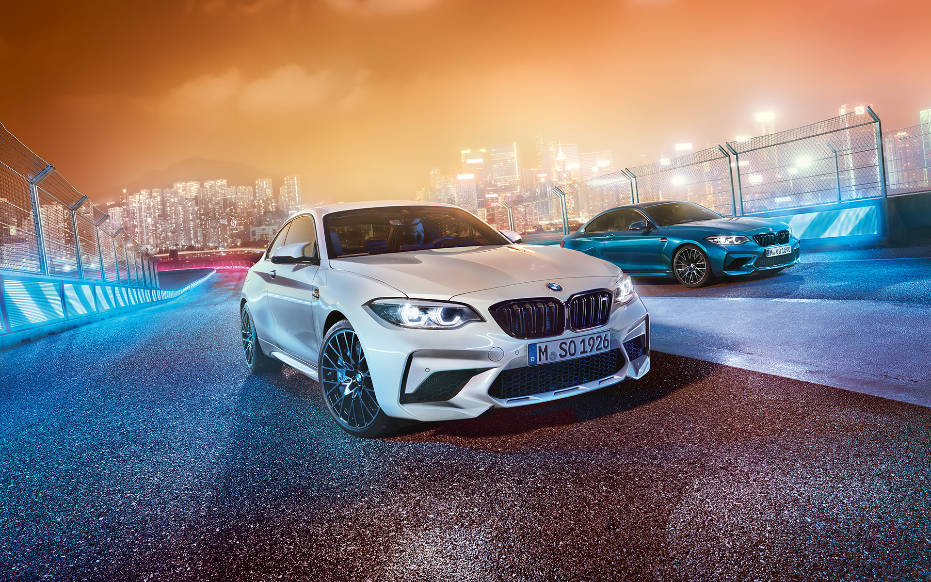 Două automobile BMW M2 Competition pe circuitul de curse Automobil BMW M2 Competition în combinaţie de alb şi albastru pe circuitul de curse în faţa unui peisaj urban nocturn.