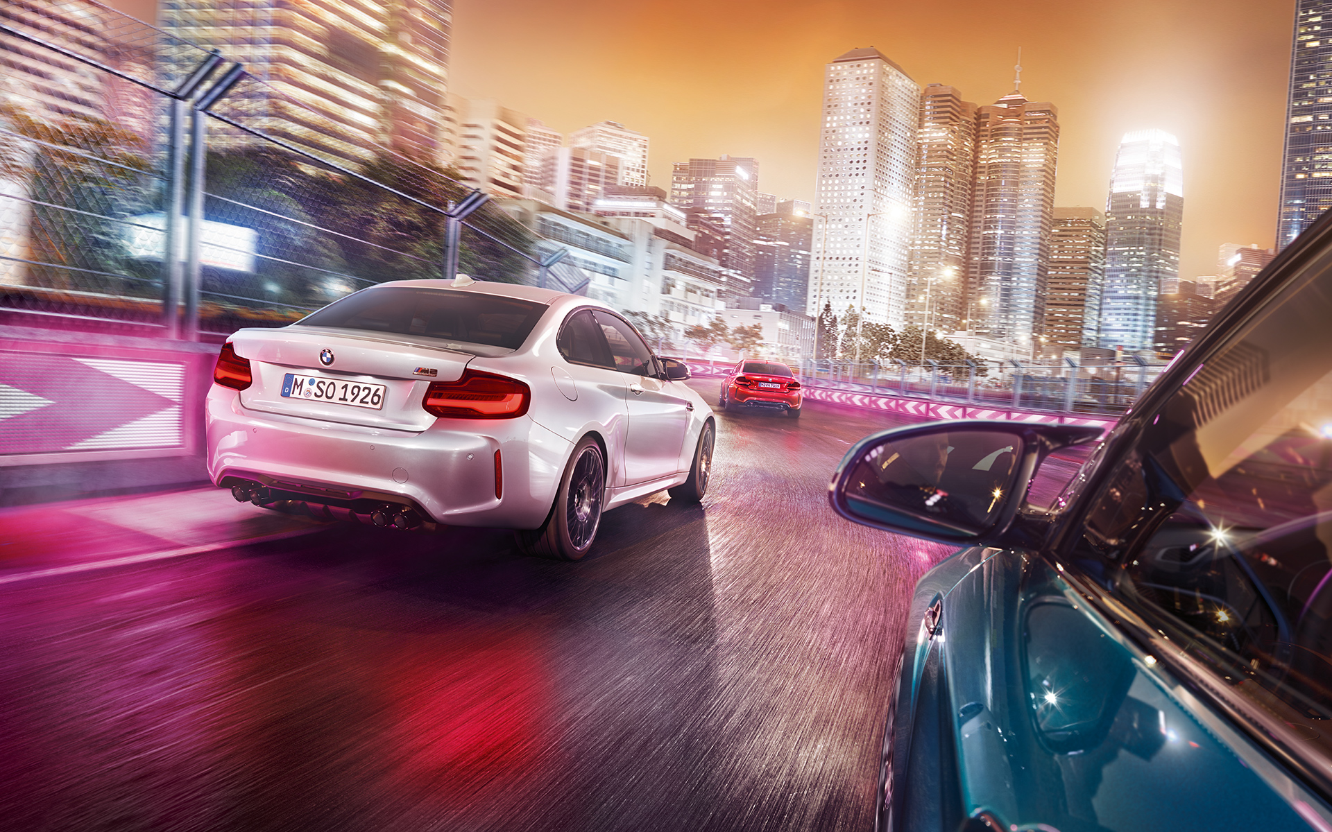 Perspectivă spate BMW M2 Competition Trei automobile BMW M2 Competition, perspectivă spate conduc pe un circuit de curse în faţa unui orizont iluminat.