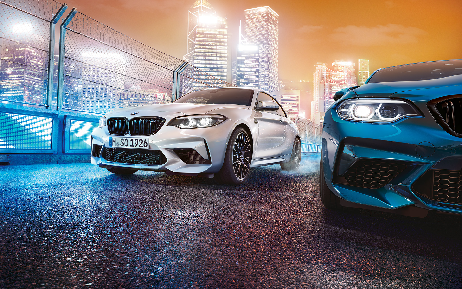 Automobil BMW M2 Competition în combinaţie de alb şi albastru, aflat în staţionare Automobil BMW M2 Competition în combinaţie de alb şi albastru cu perspectivă faţă stând în faţa unui peisaj urban nocturn.