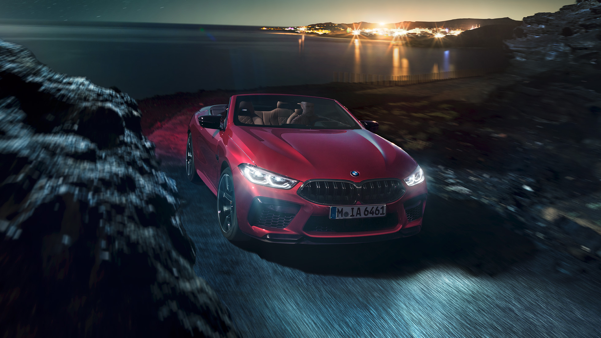 Imagine cu trei sferturi din partea frontală a BMW M8 Competition Cabriolet. Imagine cu trei sferturi din partea frontală a BMW M8 Competition Cabriolet în Roşu Motegi metalizat cu soft-top-ul deschis, rulând pe timp de noapte de-alungul coastei.