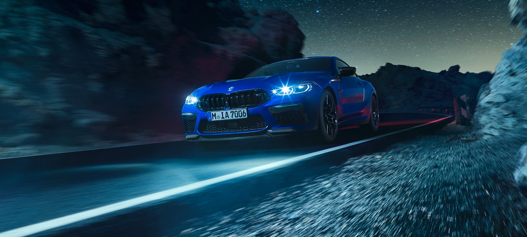 Imagine cu trei sferturi din partea frontală a BMW M8 Competition Coupé. Imagine cu trei sferturi din partea frontală a BMW M8 Competition Coupé în Albastru Marina Bay metalizat, rulând pe timp de noapte.