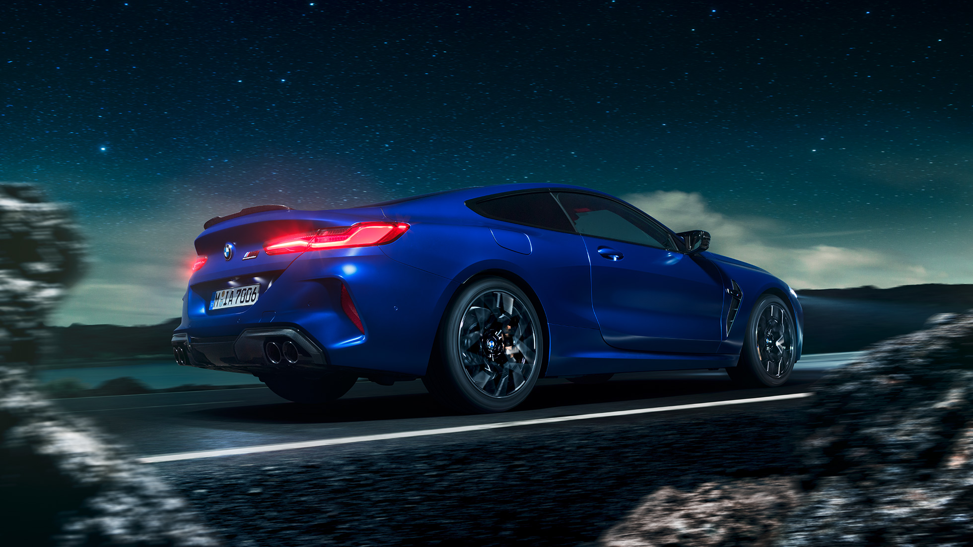 Imagine cu trei sferturi din pertea laterală a BMW M8 Competition Coupé. Imagine cu trei sferturi din partea laterală a BMW M8 Competition Coupé în Albastru Marina Bay metalizat, rulând sub cerul înstelat.