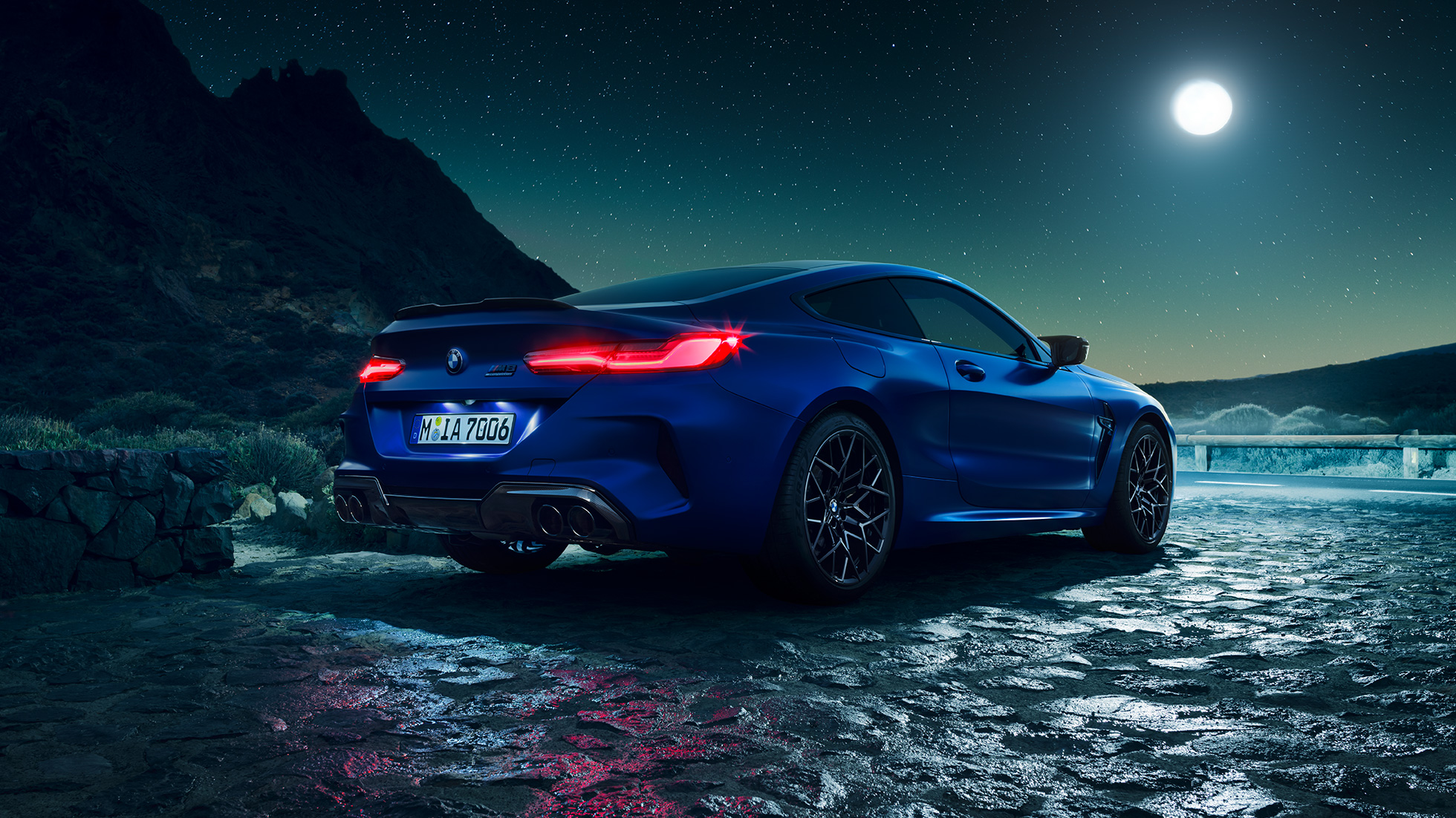 Imagine cu trei sferturi din partea spate a BMW M8 Competition Coupé. Imagine cu trei sferturi din partea spate a BMW M8 Competition Coupé în Albastru Marina Bay metalizat, cu stopuri cu tehnologie LED pornite, staţionând cu luna plină pe fundal.