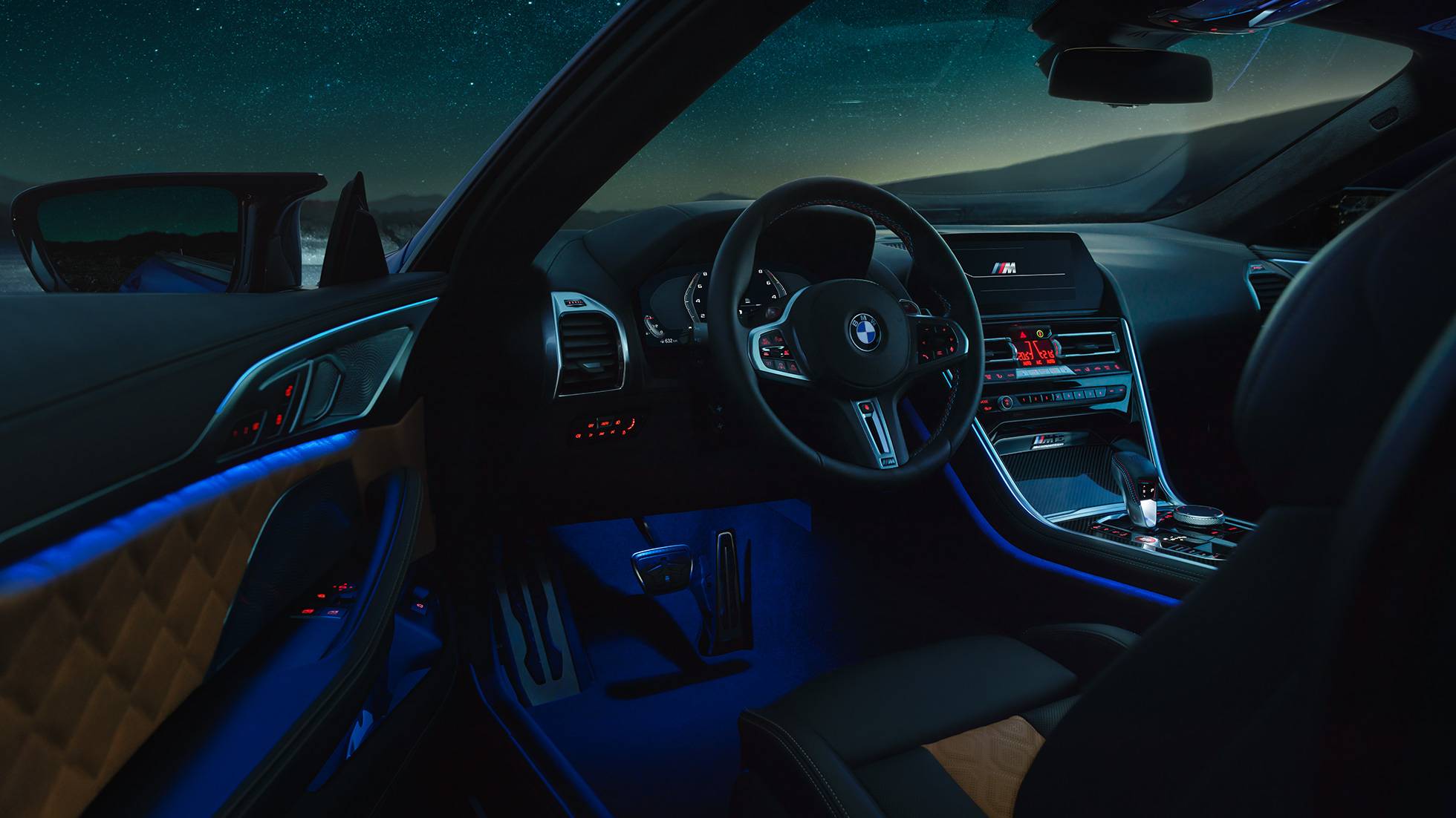 Interiorul BMW M8 Competition Coupé. Imagine cu interiorul BMW M8 Competition Coupé, cu volan M îmbrăcat în piele şi lumină ambientală albastră, pe timp de noapte.