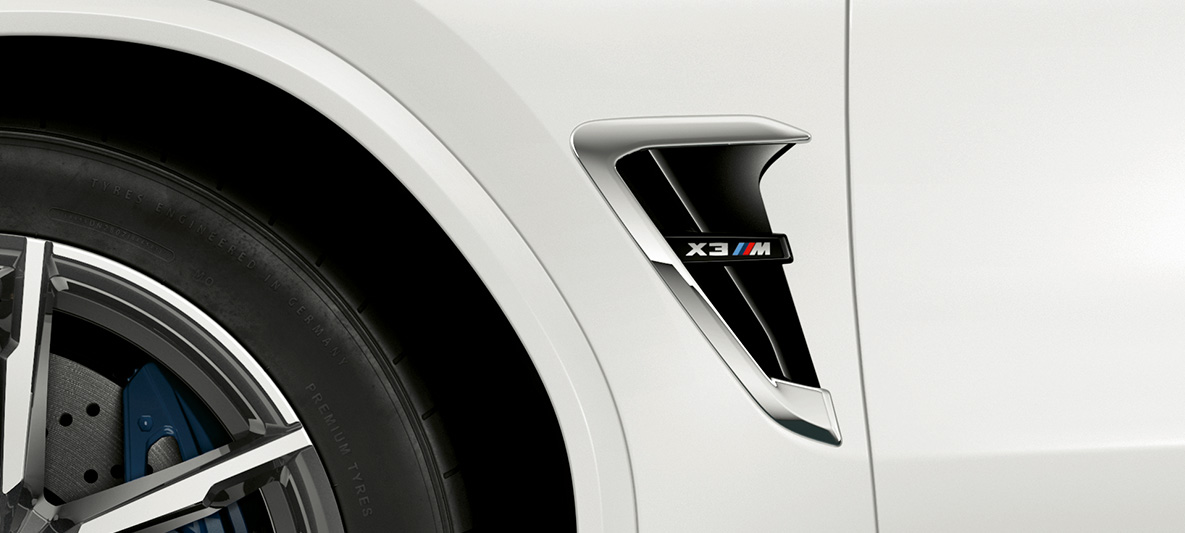 Imagine cu BMW X3 M, cu prize de aer M laterale. Imagine cu partea exterioară a BMW X3 M în Alb Alpine, cu priză de aer M laterală cu detalii cromate şi inscripţionarea denumirii modelului.