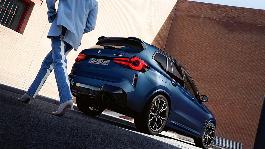 Imagine cu unghi redus cu trei sferturi din partea din spate a unui BMW X3 M Competition F97 LCI Facelift 2021, cu un model Imagine cu unghi redus cu trei sferturi din partea din spate a unui BMW X3 M Competition F97 LCI Facelift 2021 în Albastru Frozen Marina Bay, cu un model