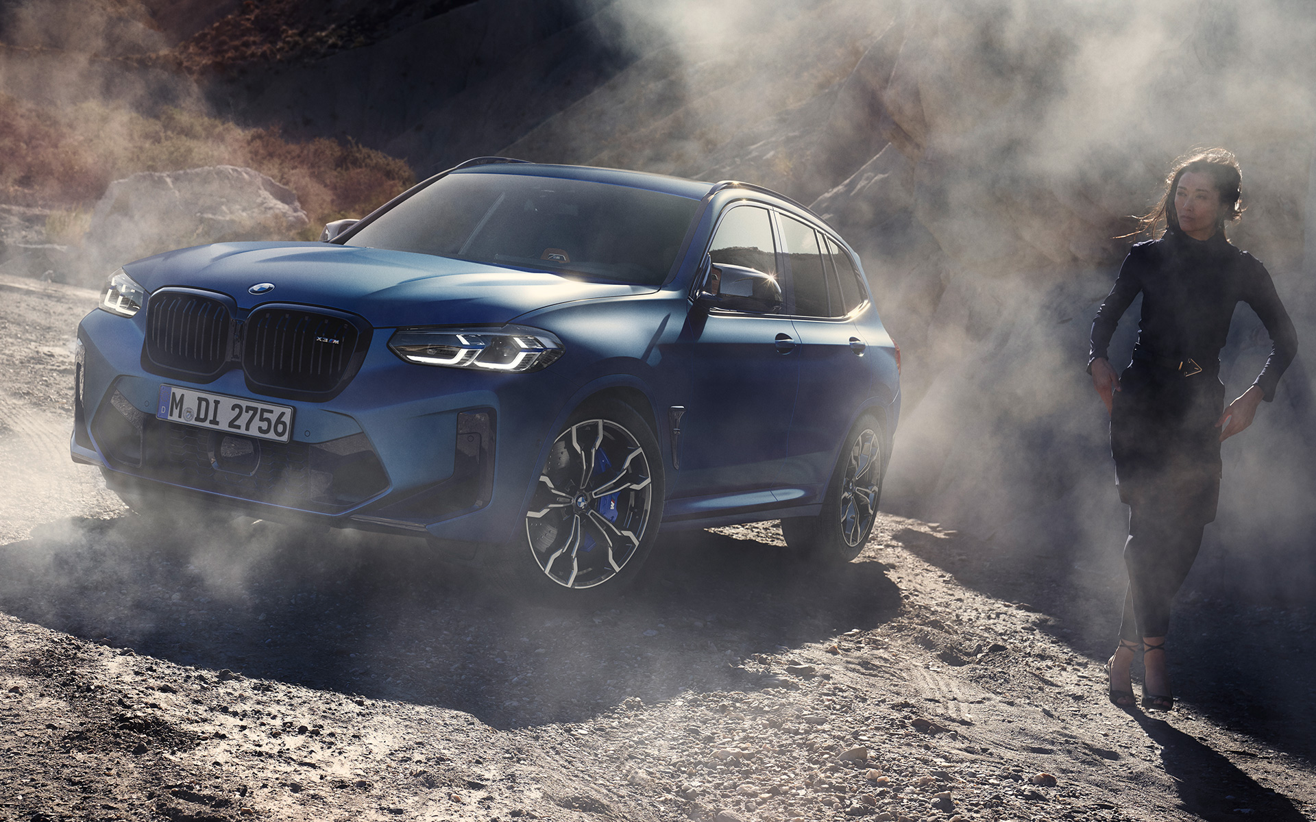 Imagine cu trei sferturi din partea frontală a BMW X3 M Competition F97 LCI Facelift 2021 în Albastru Frozen Marina Bay, staţionând pe teren stâncos cu model feminin