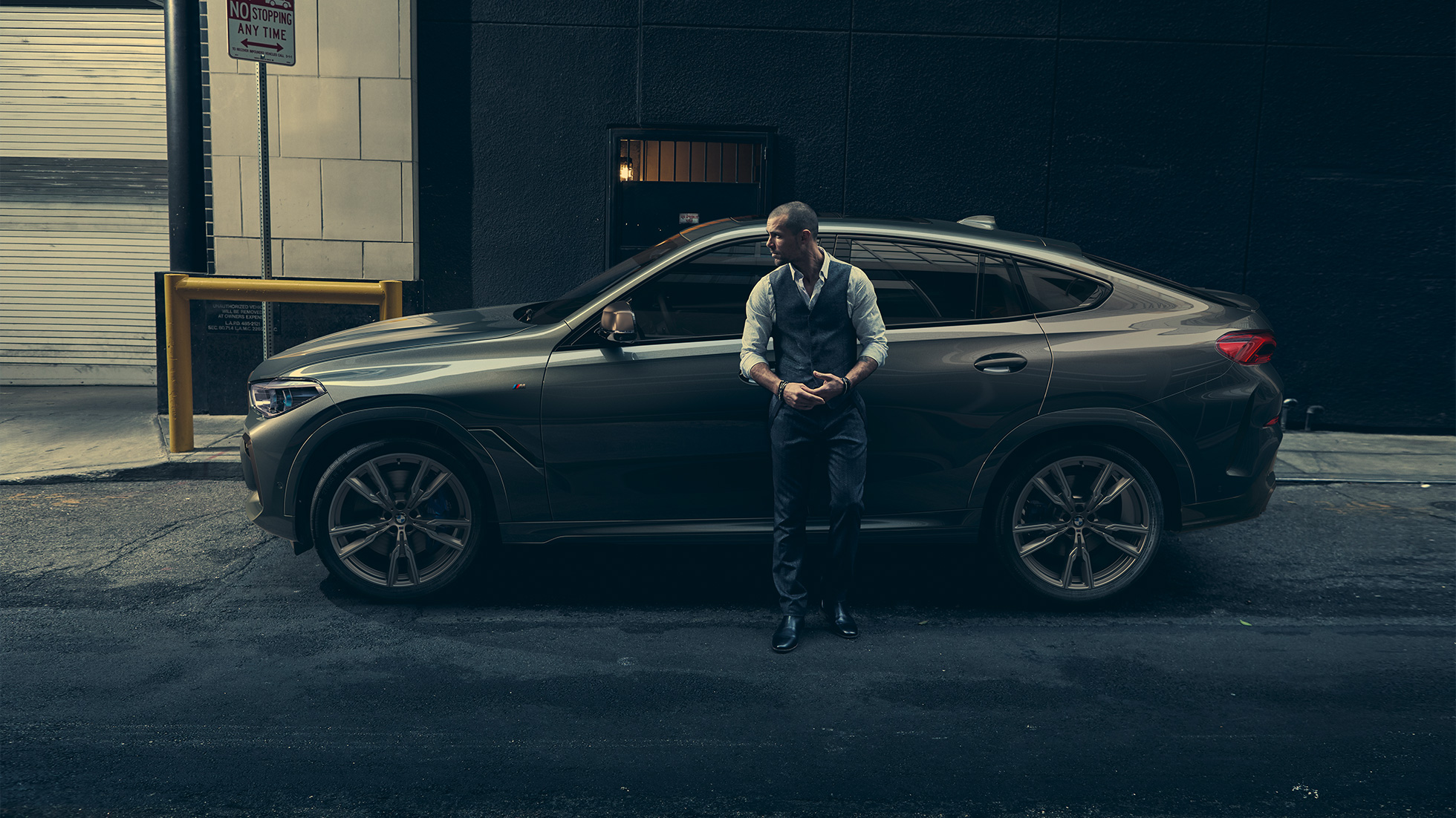 Imagine din lateral a BMW X6 M50i Imagine din lateral cu un bărbat sprijinându-se de un BMW X6 M50i în faţa unui peisaj urban