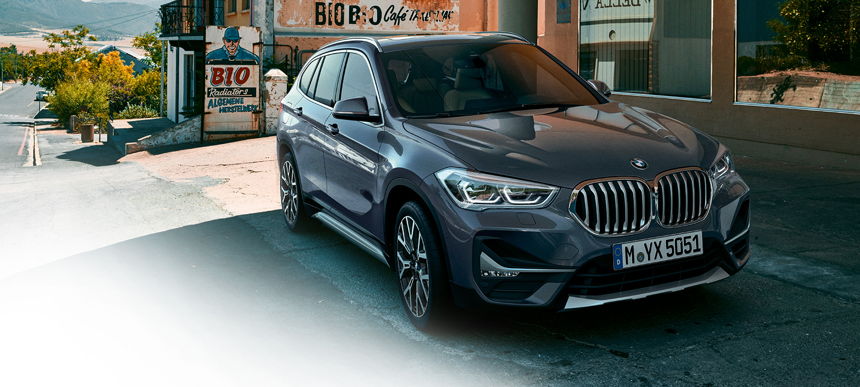 Imagine cu trei sferturi din partea frontală a BMW X1. Imagine cu trei sferturi din partea frontală a BMW X1, staţionând în faţa unui peisaj urban.