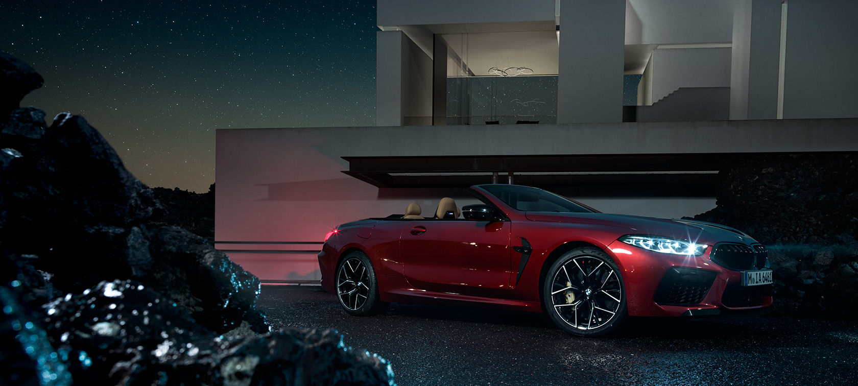 Imagine cu trei sferturi din partea laterală a BMW M8 Competition Cabriolet. Imagine cu trei sferturi din partea laterală a BMW M8 Competition Cabriolet în Roşu Motegi metalizat, staţionând pe timp de noapte în faţa unei vile.