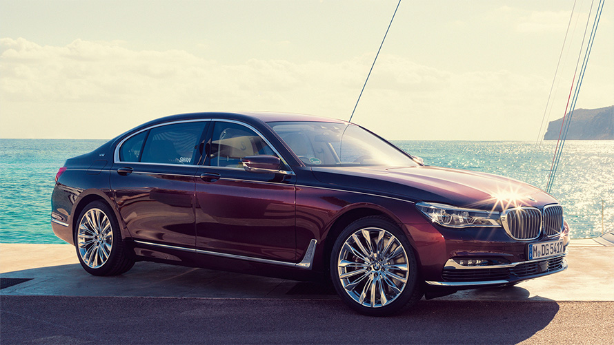 Imagine cu trei sferturi din partea laterală a BMW Individual M760Li inspirat de Nautor’s Swan G12 2017, staţionând la debarcader în faţa unui iaht cu vele în golf